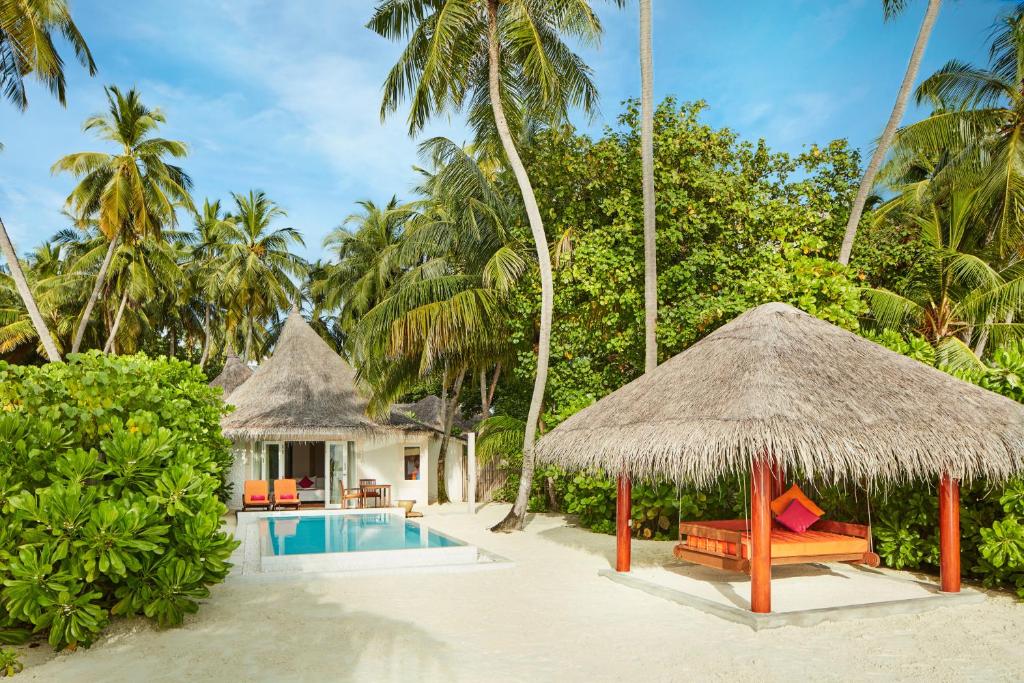 A serene outdoor area features a private pool surrounded by lush greenery. A traditional thatched-roof pavilion offers seating with vibrant cushions. The villa is set against a backdrop of swaying palms, highlighting a tranquil beachside atmosphere.