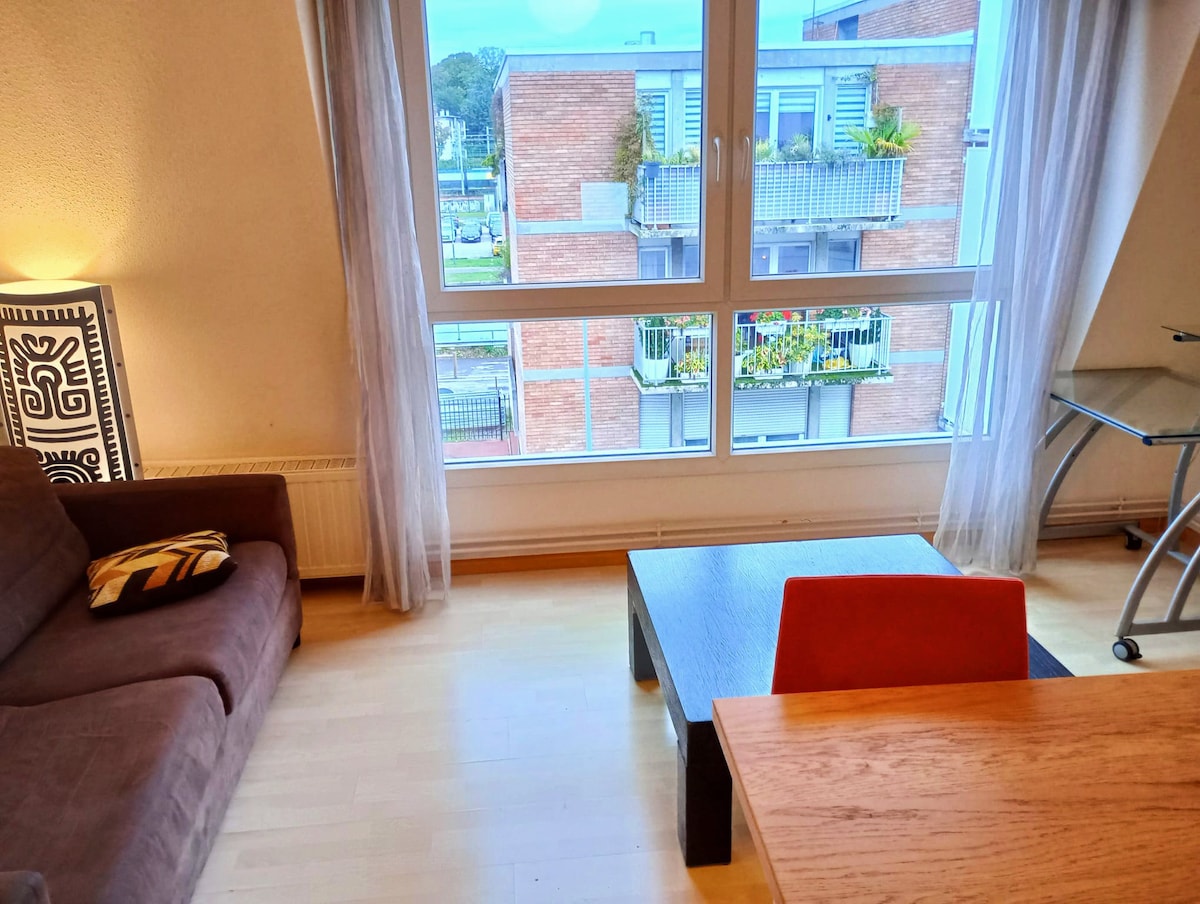 A living area features a brown sofa with a decorative cushion, accompanied by a simple wooden coffee table. Natural light enters through large windows, revealing a view of a neighboring building with greenery on the balcony. A small dining table with an orange chair is also present.