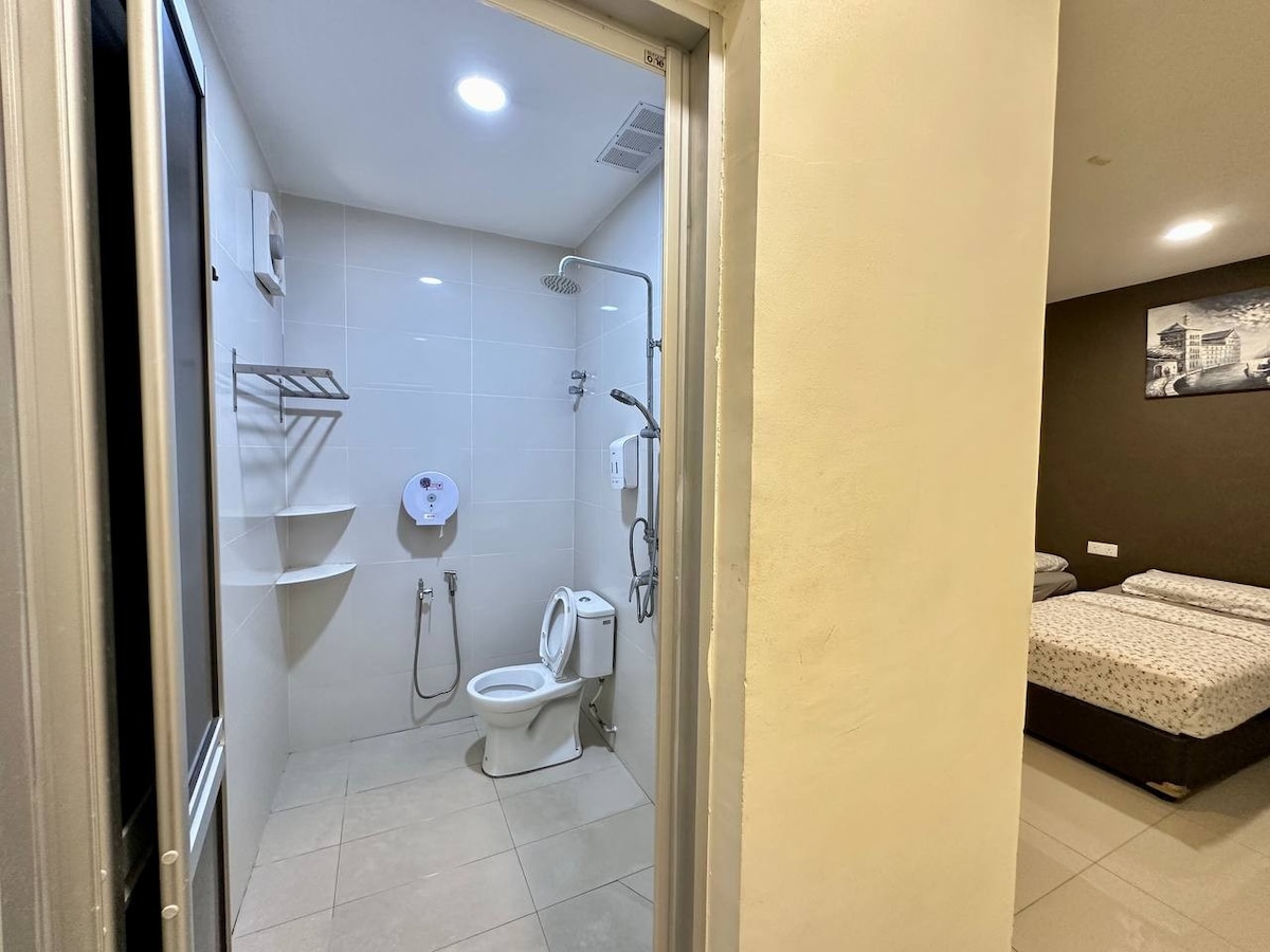 A modern bathroom is featured, equipped with a shower fixture, wall-mounted storage shelves, and a toilet. Natural light from overhead lighting enhances the clean tiled surfaces. The adjoining bedroom is partially visible, showcasing a laid bed with decorative bedding.