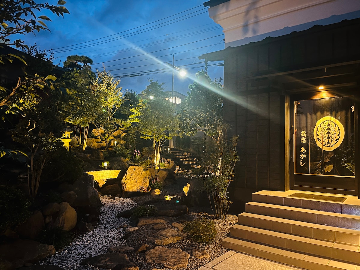 Evening light casts a soft glow over a landscaped garden featuring natural stone pathways and thoughtfully placed greenery. A traditional Japanese lantern is visible, and the entrance to the building is highlighted, showcasing the detailed craftsmanship of the architecture.