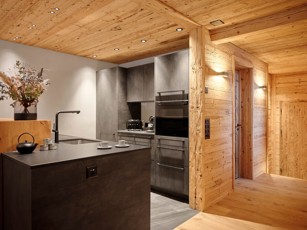 A modern kitchen features sleek gray cabinetry complemented by wood accents. An island with a minimal design sits in the foreground, presenting a stovetop and seating area. Natural light enhances the warm wooden ceiling, creating a seamless connection with the surrounding interior.