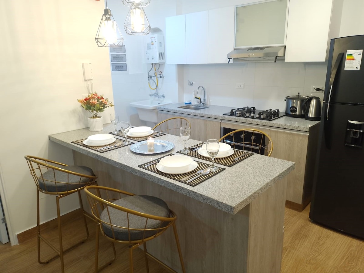 The kitchenette features a modern granite countertop with three high-backed chairs arranged around it. Essential kitchen appliances are visible, including a stovetop and refrigerator. Table settings include plates and glasses, complemented by a small flower arrangement adding a touch of color.