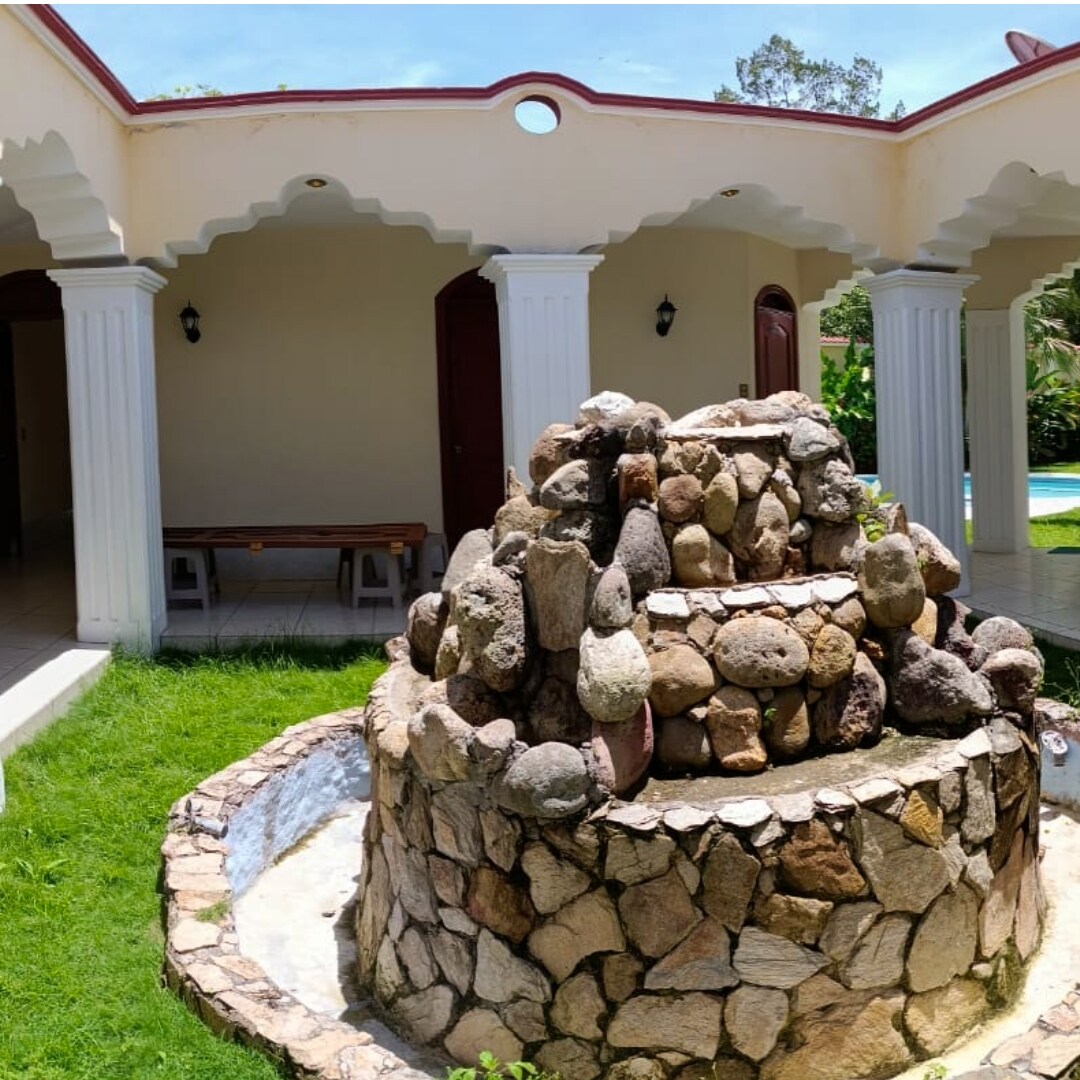 A stone fountain is centrally positioned, featuring a layered design crafted from various natural rocks. Lush greenery surrounds the fountain, while the shaded area is highlighted by nearby columns and a wooden table, creating a serene outdoor space.