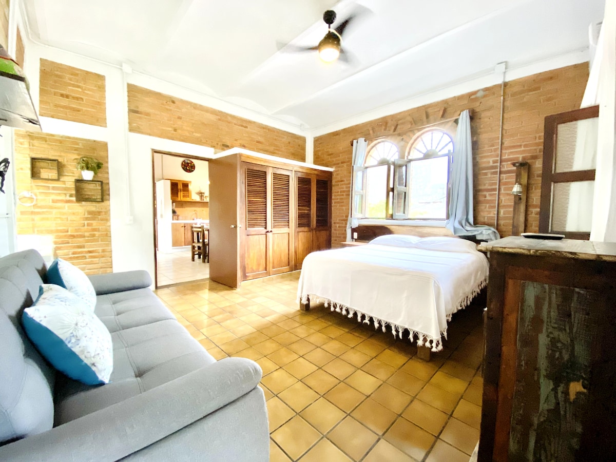 The studio features a comfortable sleeping area with a spacious bed dressed in white linens. A cozy seating area is provided by a light grey sofa, while warm brick walls add character. Natural light fills the room through an arched window, enhancing the inviting atmosphere.
