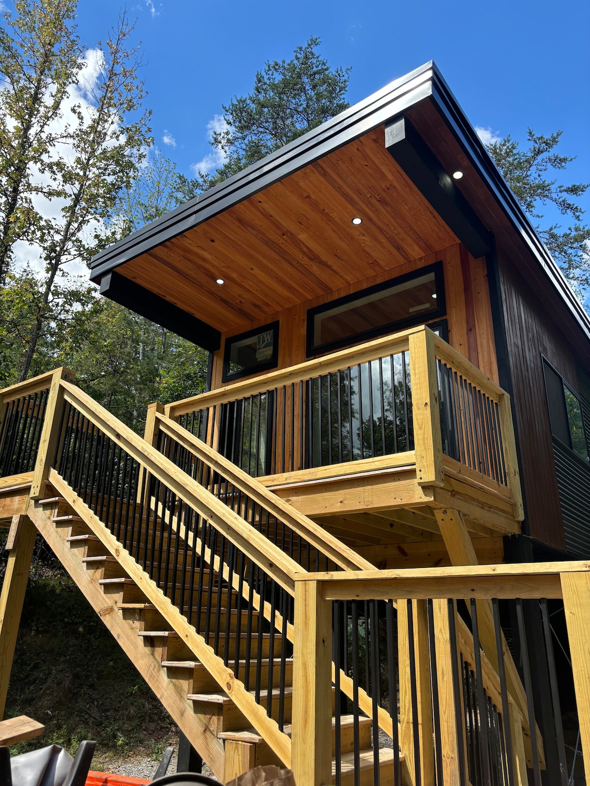 The treehouse exterior features a contemporary design with a wood and metal facade. A sturdy wooden staircase leads to the entrance, framed by black railings. Tall windows invite natural light, and the roof is highlighted by sleek overhangs, enhancing the modern aesthetic.