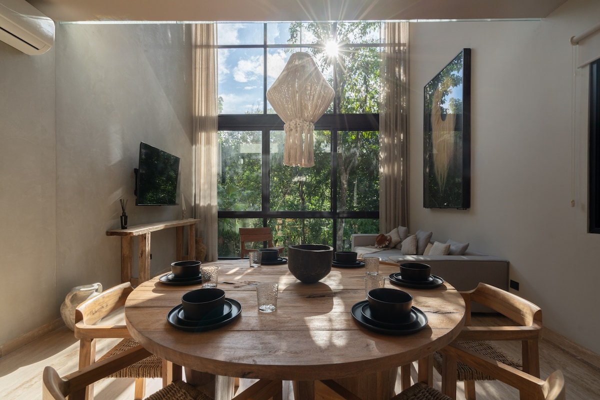 A bright open-concept dining area features a round wooden table set for six. Natural light floods the space through large windows, highlighting the surrounding greenery. A woven light fixture hangs above, and a comfortable seating area is visible in the background.