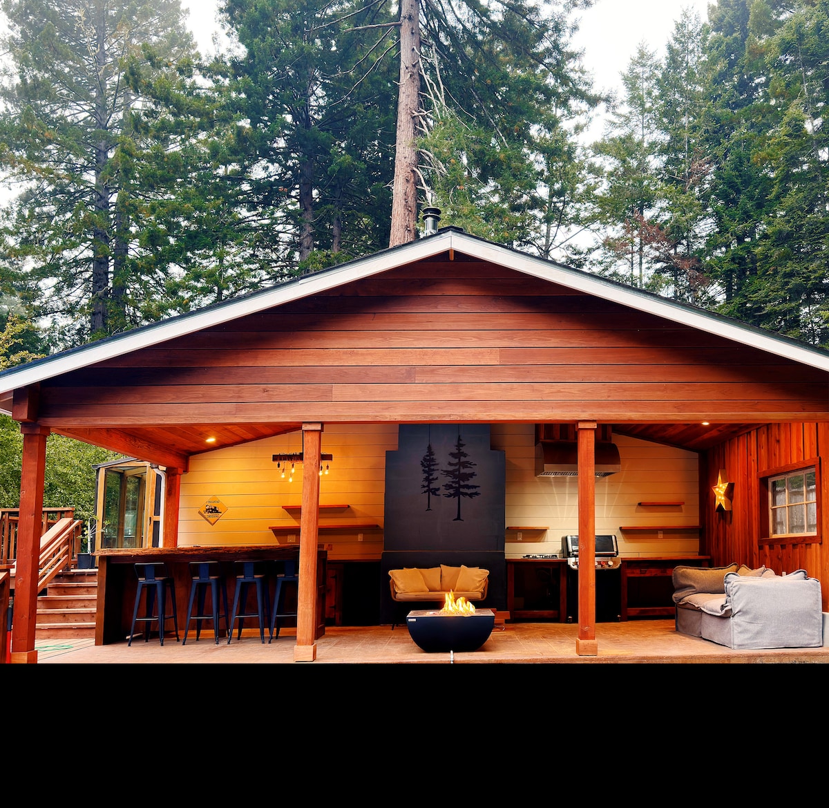 A spacious outdoor veranda is showcased, featuring a modern seating area with a fire pit at the center. The rustic wooden structure is framed by towering redwoods, creating a serene backdrop for relaxation and social gatherings.
