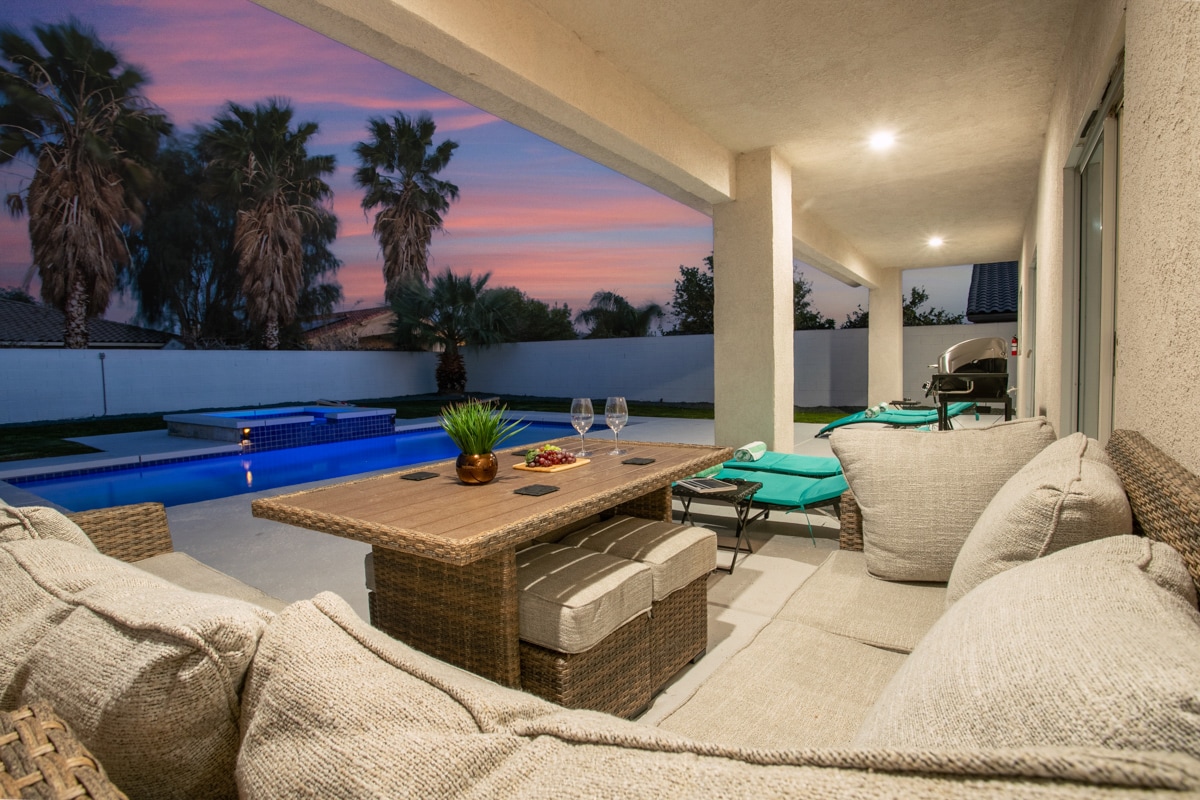 An outdoor seating area features a large woven sofa and a coffee table, set against a backdrop of a swimming pool and palm trees. Soft lighting highlights the space as sunset colors paint the sky, creating a tranquil atmosphere for relaxation.