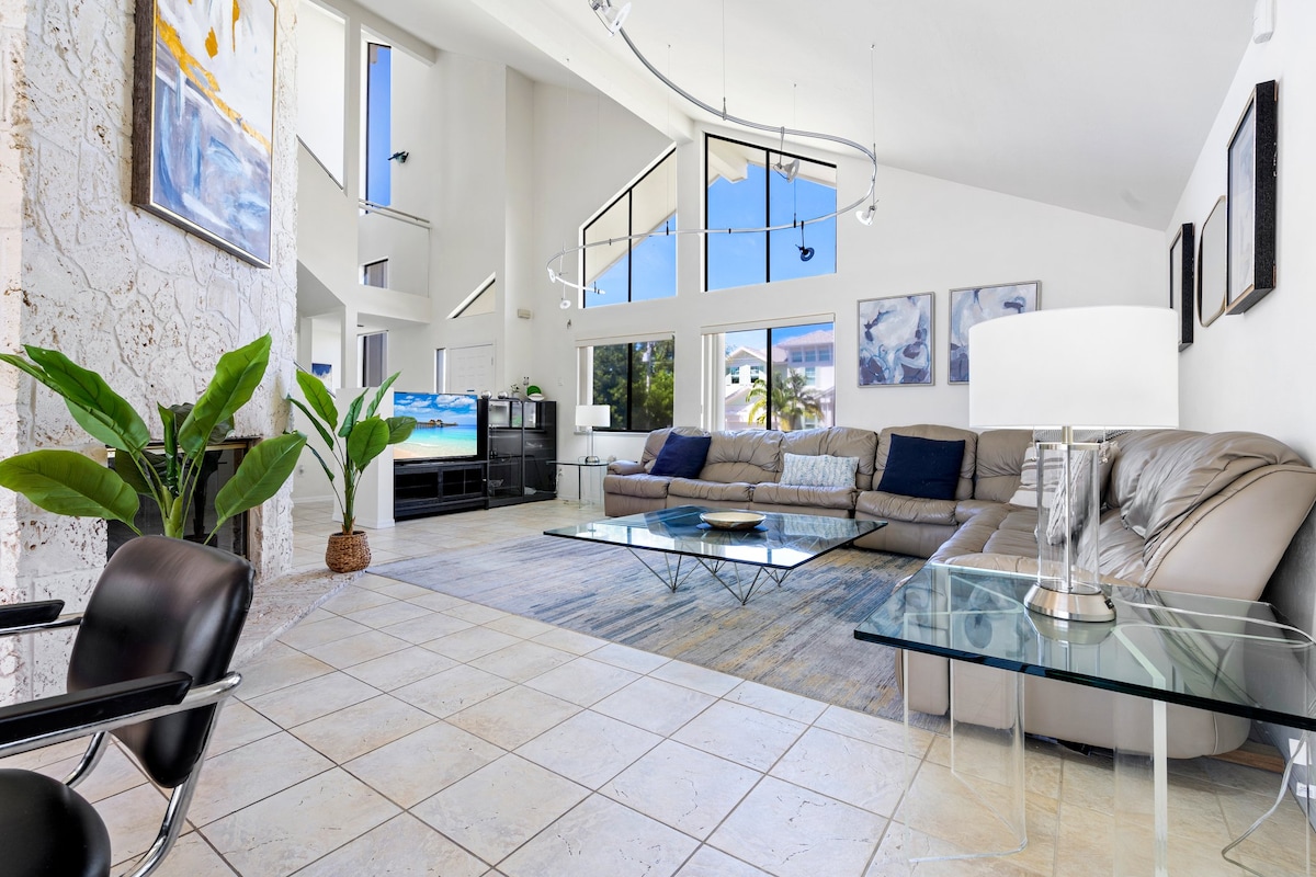 A spacious living area is illuminated by natural light streaming through large windows. A comfortable sectional couch surrounds a glass coffee table, complemented by a decorative rug. A television is positioned against the wall, and vibrant green plants add a touch of nature to the space.