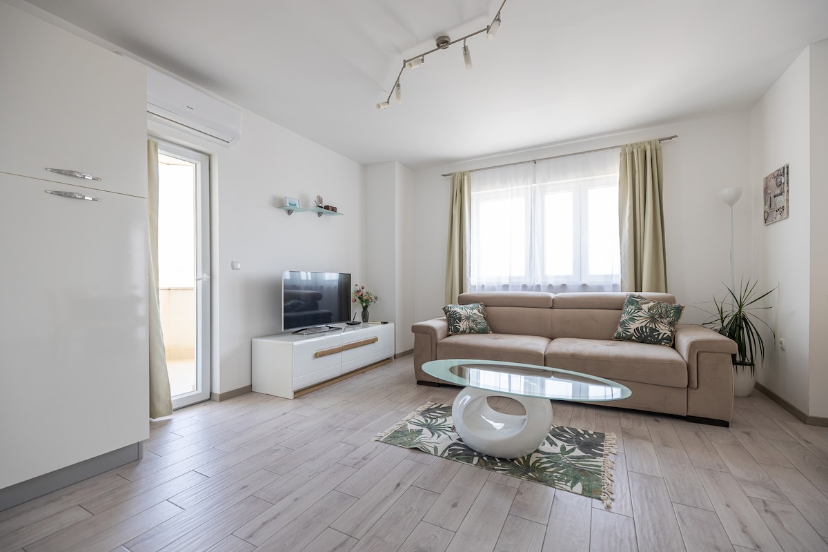 A modern living room features a comfortable sofa adorned with decorative cushions. A sleek glass coffee table sits in the center, while a television is mounted on the wall opposite. Natural light fills the space through large windows, complemented by airy curtains.