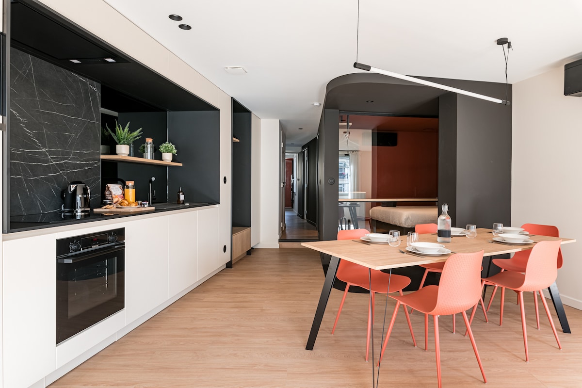A modern open kitchen and dining area features sleek black cabinetry and a light-colored countertop. A wooden dining table is surrounded by coral-colored chairs, set for a meal. Natural light fills the space, highlighting the minimalist design and thoughtful layout.