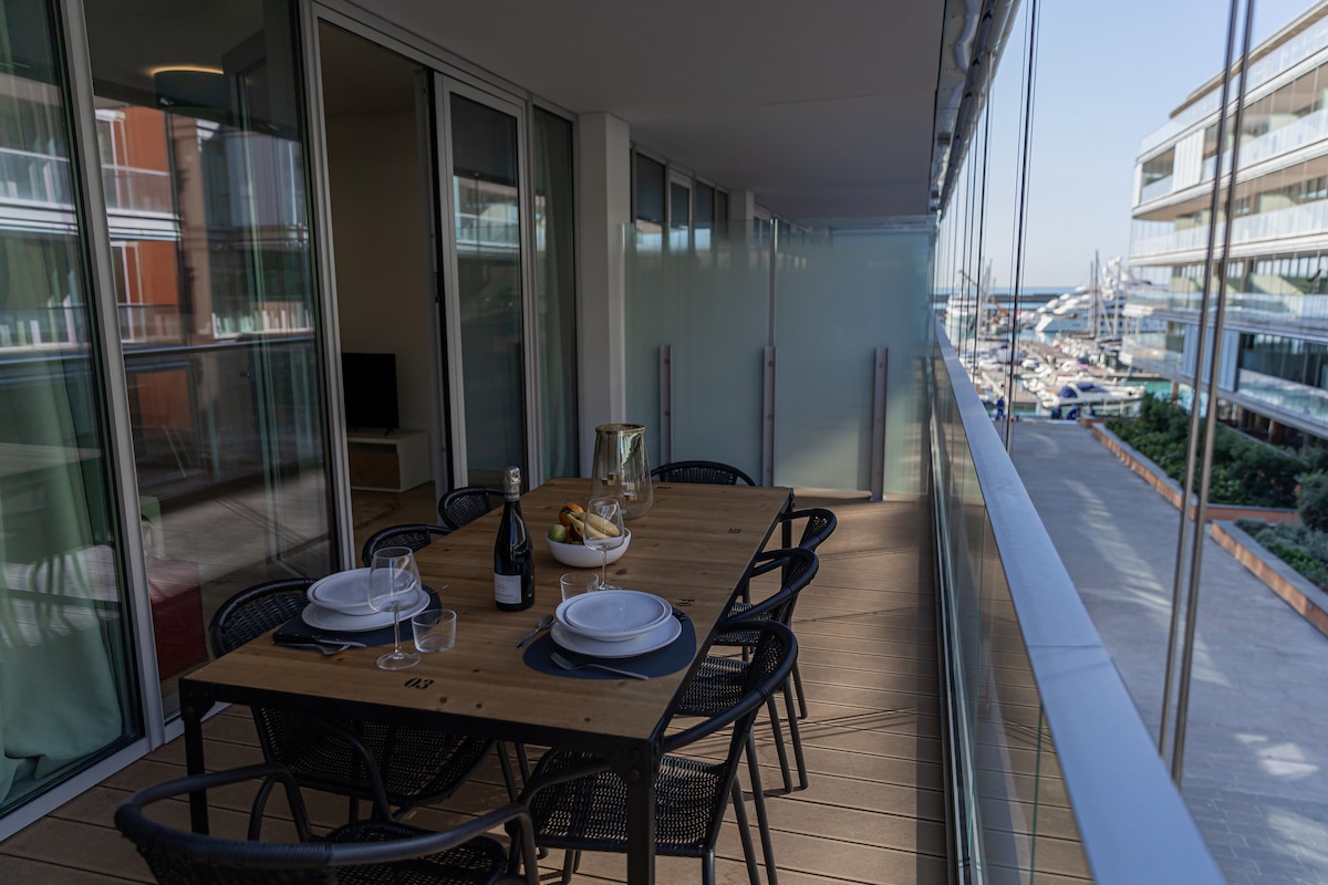 An outdoor dining area features a wooden table set for six, accompanied by black woven chairs. A bottle of wine and a fruit arrangement are positioned on the table. Floor-to-ceiling glass panels provide views of the marina and surrounding waterfront development.