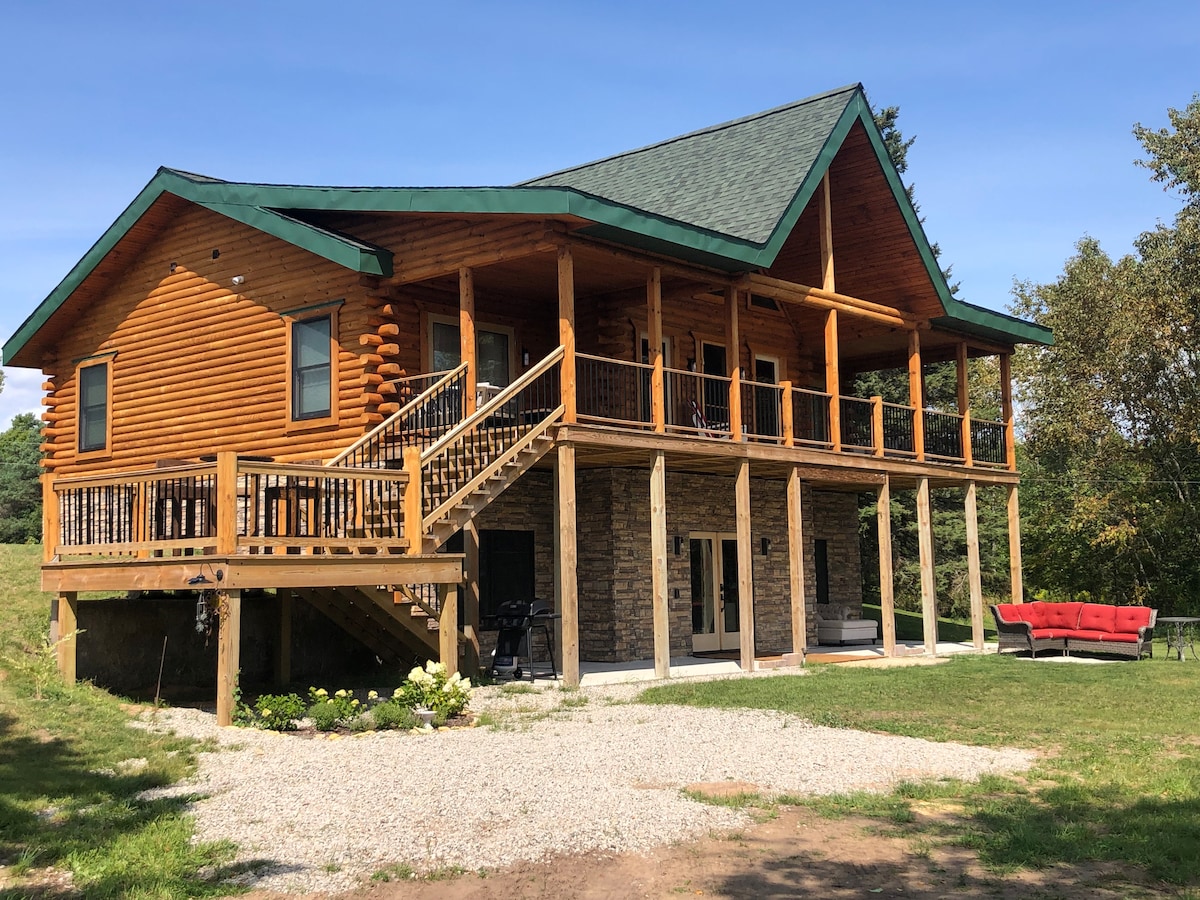 A charming log cabin is showcased, featuring a two-level structure with a sloped green roof. The expansive front deck is supported by sturdy columns and provides access to the upper level, while a lower patio area is visible. A red outdoor sofa adds a pop of color to the surrounding greenery.