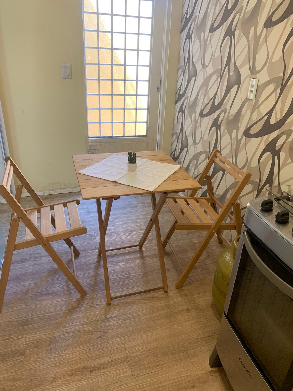 A small dining area features a light wood table surrounded by two folding chairs. A simple tablecloth covers the table, which holds a set of salt and pepper shakers. Natural light filters through a nearby glass door, creating a warm and open feel.