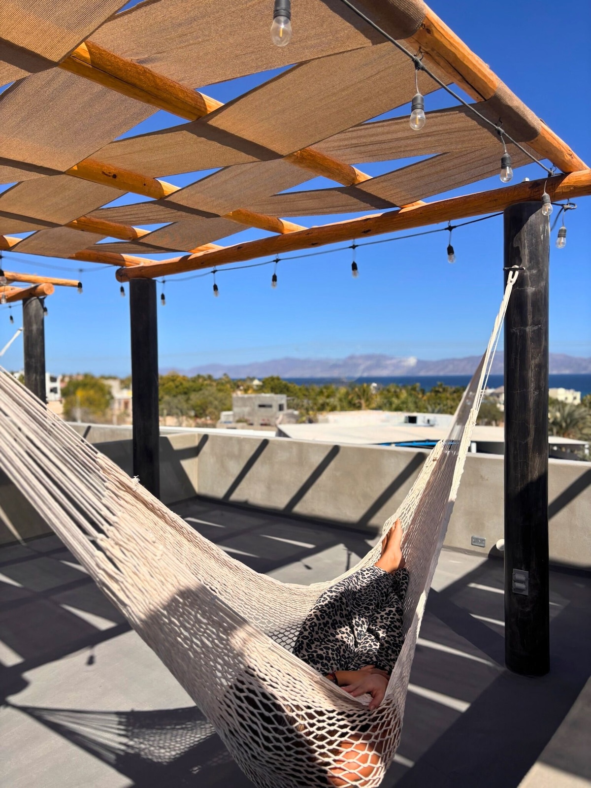A relaxing rooftop area features a spacious hammock suspended beneath a wooden pergola. Decorative lights hang from the structure, enhancing the atmosphere. In the background, mountains and a clear blue sky create a serene setting, perfect for unwinding and enjoying the views.