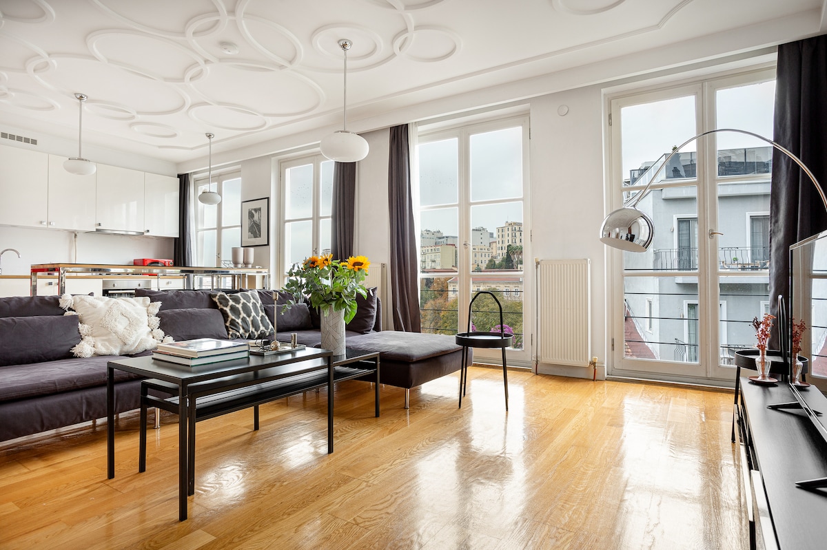 The spacious living area features large windows that allow natural light to fill the room. A stylish sofa is complemented by a black coffee table, while a vase with colorful flowers adds a touch of life. A modern floor lamp stands nearby, enhancing the comfortable setting.