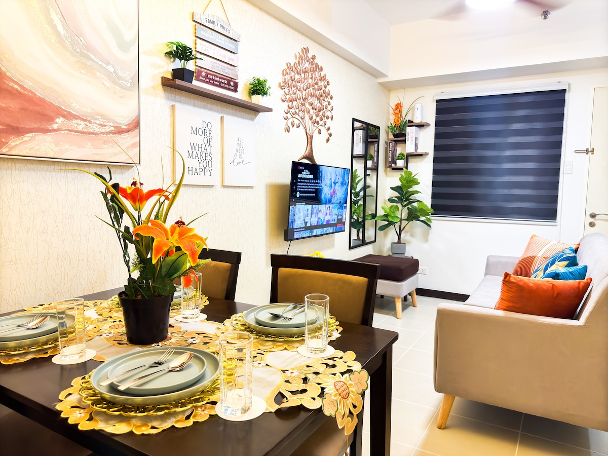 A modern living and dining area features a black dining table set for four, adorned with floral placemats and glassware. A three-seater sofa adds comfort, while decorative wall shelves display potted plants and artwork. A flat-screen TV is mounted on the wall.