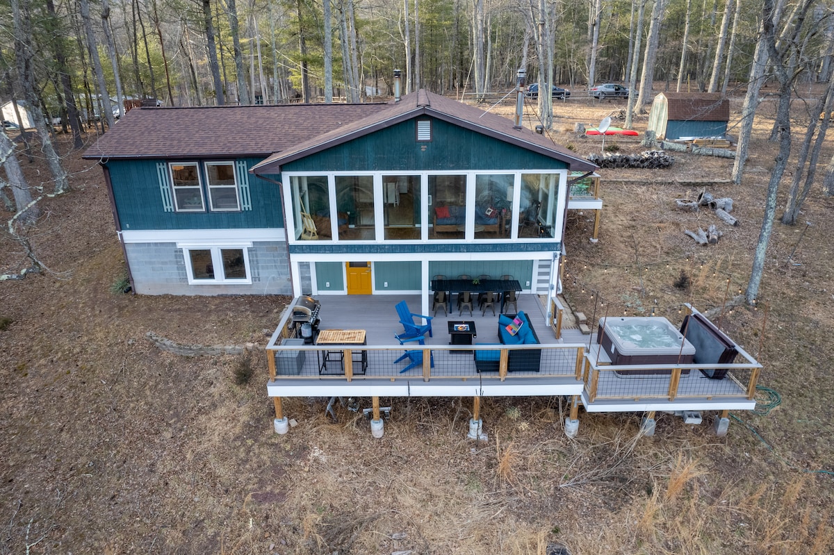 An aerial view showcases the cabin nestled in a wooded area, highlighting its multi-level structure. The spacious deck features a hot tub, a grill, and outdoor seating. The surrounding landscape is dotted with trees, enhancing the natural setting.