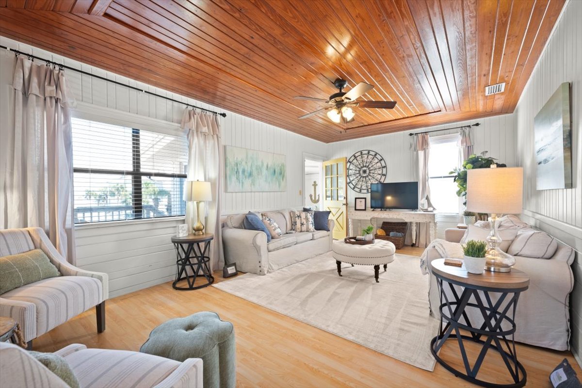 A bright living room features wooden ceiling panels, large windows allowing natural light, and a mix of comfortable seating options. A round ottoman sits in the center, surrounded by decorative tables and soft furnishings. Wall art contributes to the inviting ambiance of the space.