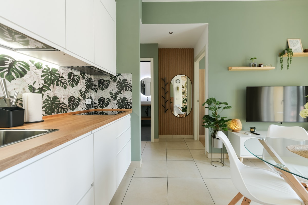 A modern kitchen area is visible, featuring white cabinetry and a wooden countertop. A decorative backsplash with botanical patterns adds texture. The open floor plan leads to a cozy dining space, showcasing a round table and stylish chairs. A large mirror and greenery provide warmth to the environment.