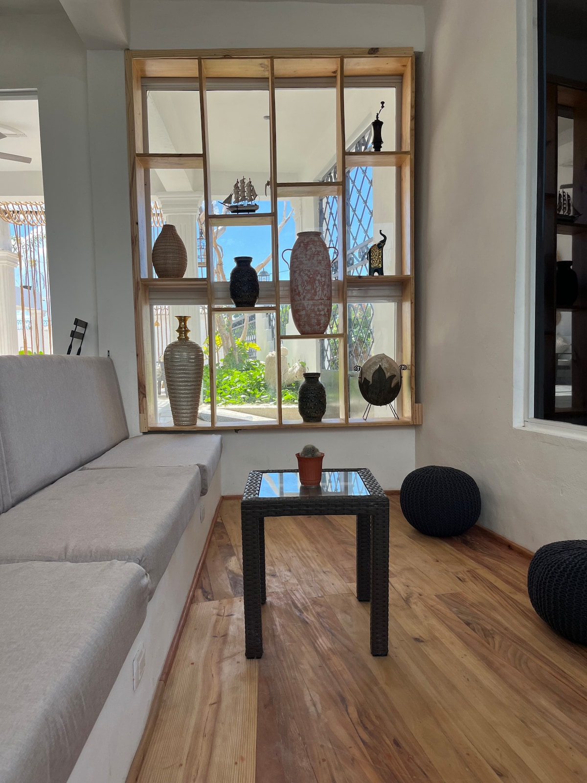 A bright seating area features a minimalist design with a light-colored sofa and a small glass-top table. Decorative objects, including vases and sculptures, are displayed within a wooden-framed window structure, allowing natural light to illuminate the space. A view of greenery can be seen in the backdrop.
