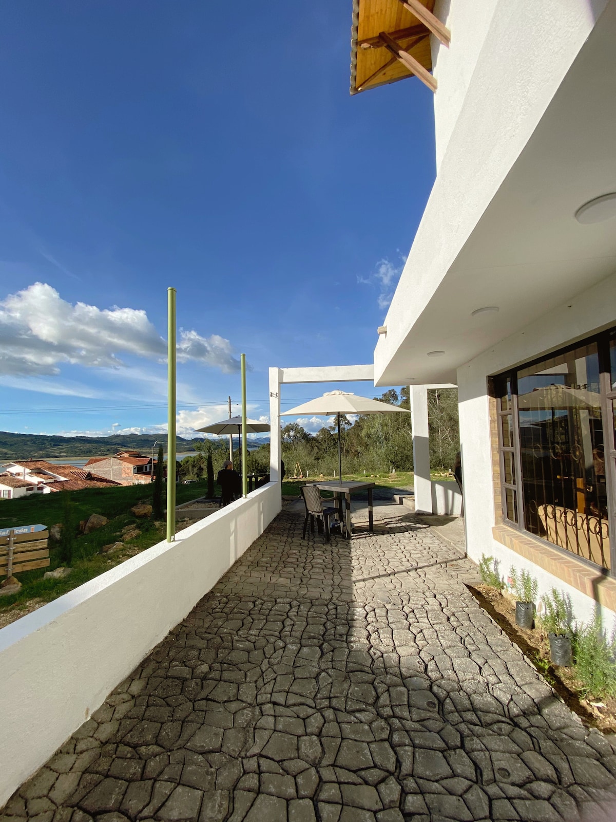 An outdoor terrace is depicted, featuring a large table covered with an umbrella. The stone pathway leads to a lush green landscape, complemented by distant mountains under a clear blue sky. Views of the nearby town are visible from the terrace.