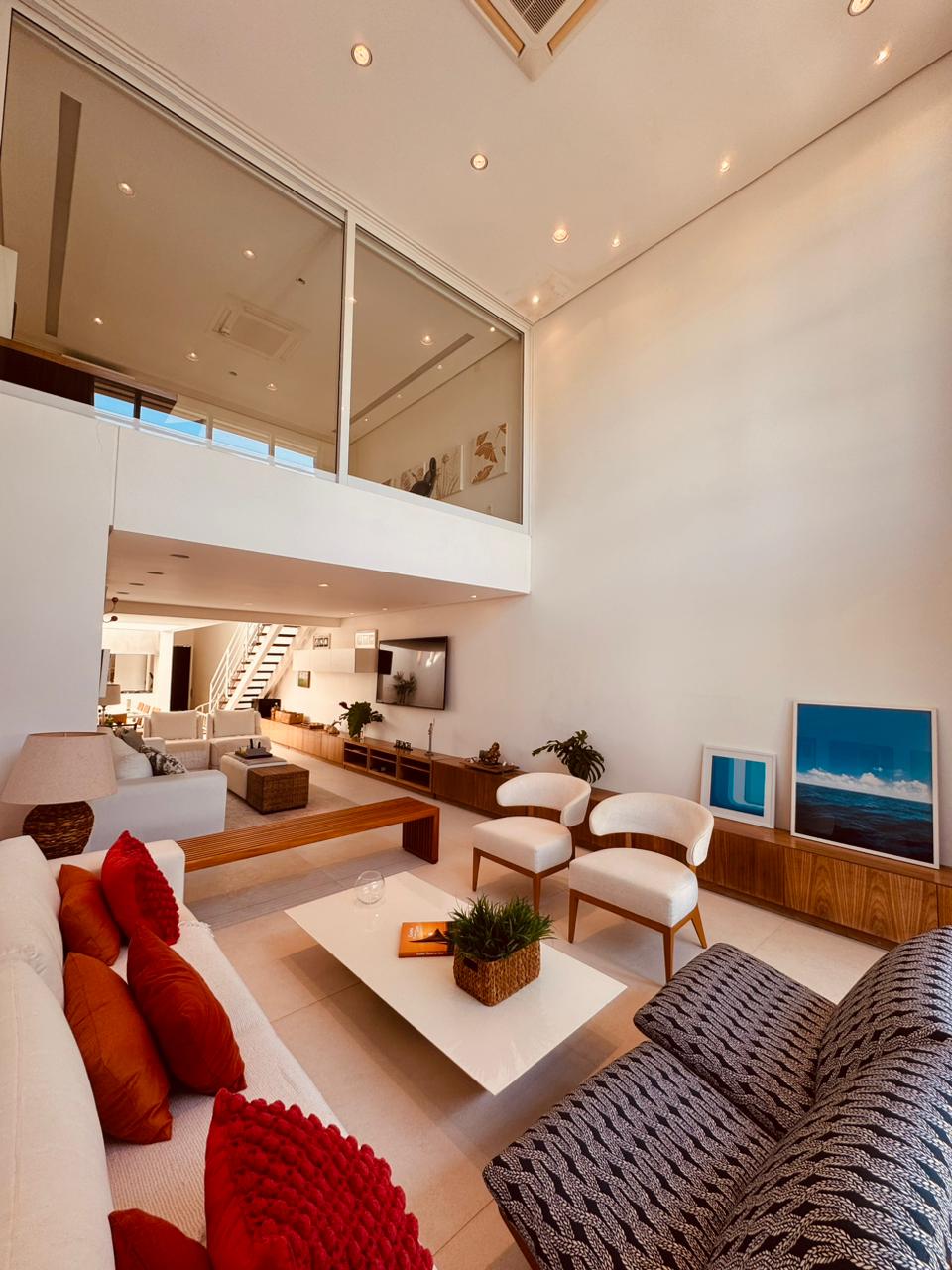 A bright living area features high ceilings and large windows that allow natural light to fill the space. A comfortable sofa adorned with decorative cushions faces a sleek coffee table, while minimalist furniture lines the walls. A staircase leads to another level, enhancing the open layout.