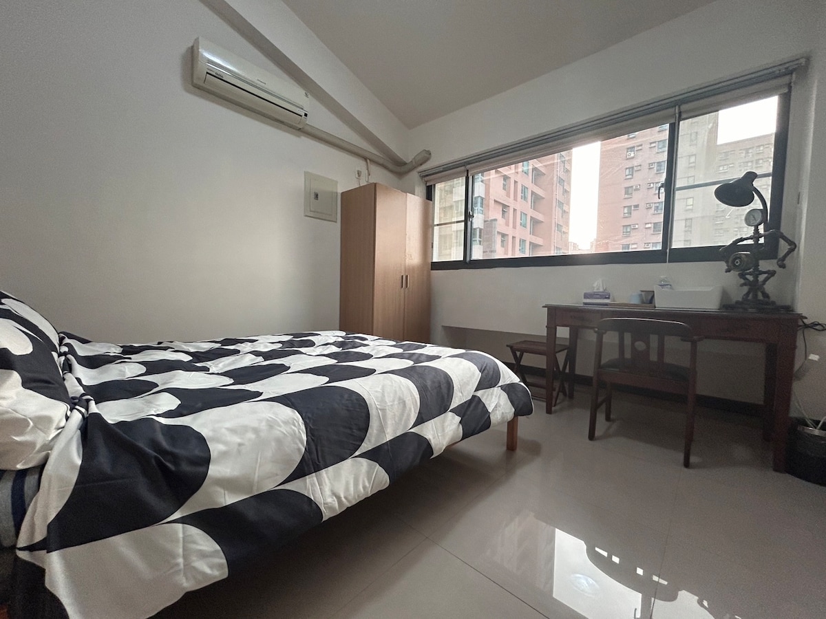 A well-lit bedroom features a spacious bed with a black and white polka dot duvet. Natural light enters through large windows, illuminating a wooden desk and chair set against the wall. A closet stands nearby, and an air conditioning unit is mounted above.