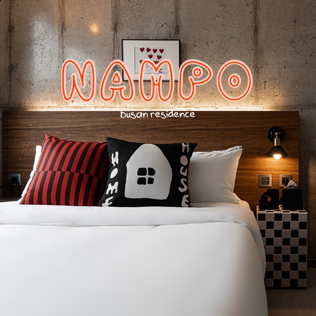 A cozy sleeping area features a queen bed with crisp white linens and distinctive black-and-white themed pillows. A wooden headboard is complemented by modern bedside lamps, while a neon sign adds a pop of color to the industrial-style walls.