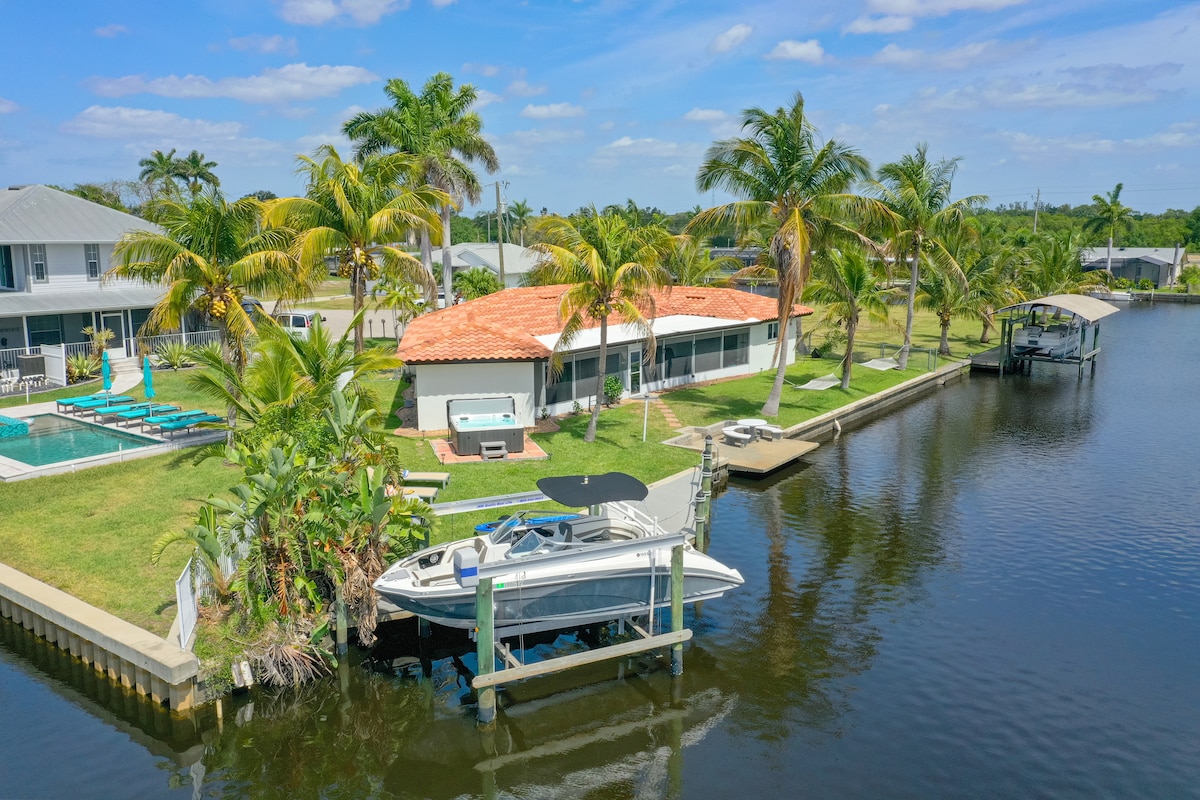 A waterfront view highlights the property surrounded by palm trees. A boat is docked along the water, while lounge chairs are visible by a pool in the yard. The home features a red tile roof, and nearby structures are seen across the water.