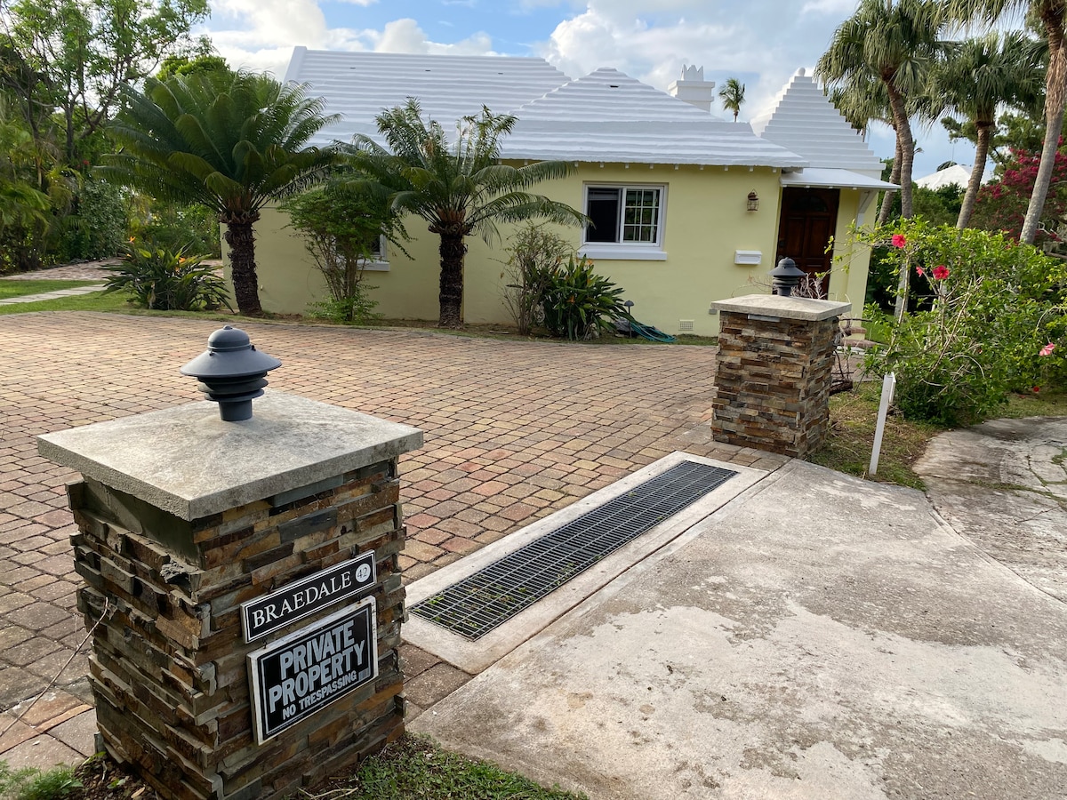 The exterior of Braedale is presented, featuring a charming stand-alone house surrounded by lush greenery. A stonewall entrance marks the property, with a sign indicating private access. The roof is light-colored, and the driveway is paved, leading to the welcoming front door.