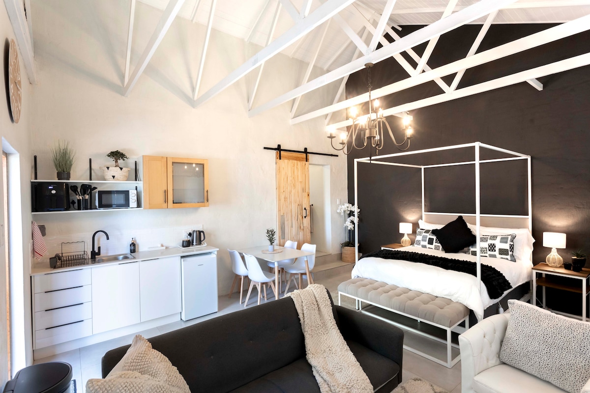 An open plan interior space features high ceilings with exposed beams. A four poster queen bed is positioned next to a minimalist nightstand. A dining area with a four-seater table is visible, along with a well-equipped kitchen showcasing modern appliances, and a cozy seating arrangement.
