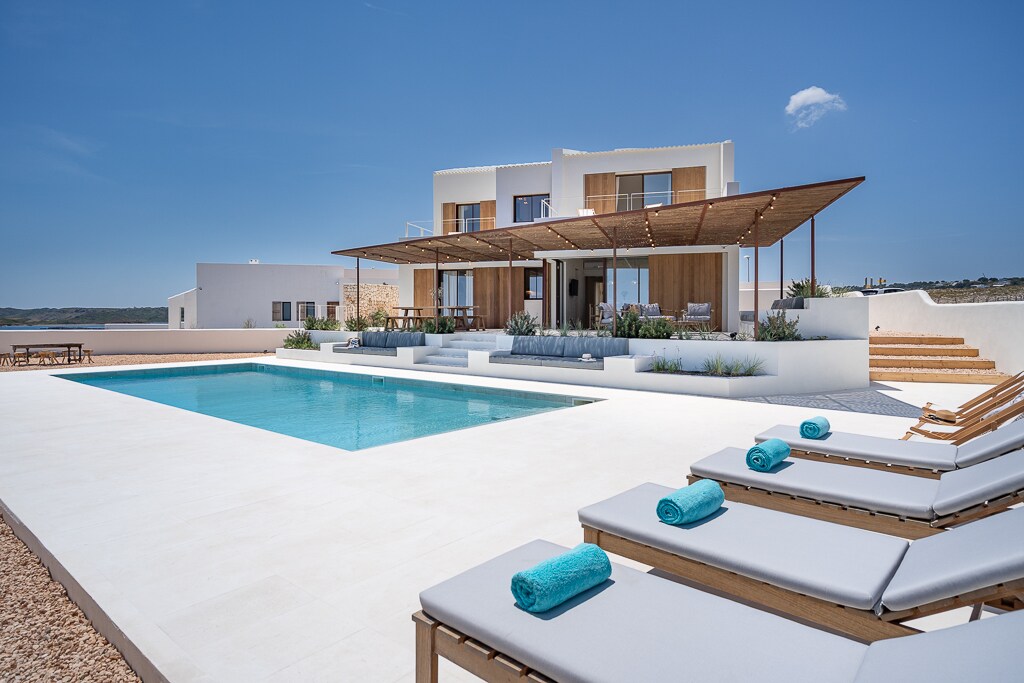 The modern villa features a large swimming pool surrounded by comfortable sun loungers. A spacious terrace with a wooden awning provides shade for outdoor dining. Expansive glass doors lead to the interior, revealing a bright and inviting atmosphere. Scenic views of the landscape enhance the outdoor experience.