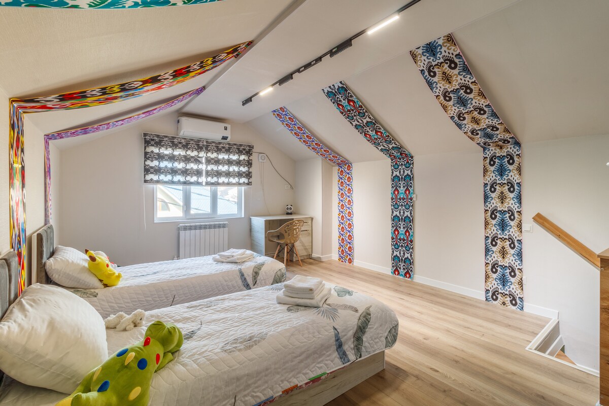 A bright and spacious room features two beds positioned apart, with colorful decor accents along the walls. A work desk with a chair is placed near a window. Natural light filters through the textile-patterned window treatments, enhancing the inviting atmosphere of the space.