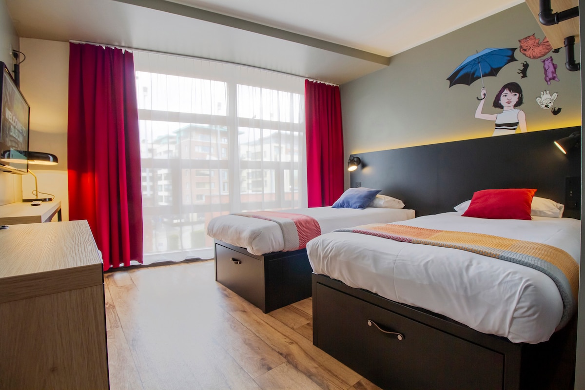 A bright room features two single beds with colorful throw pillows. A wooden dresser and a wall-mounted TV are positioned opposite the beds. Large windows draped with red curtains allow ample natural light, while a unique wall mural adds a playful touch to the space.