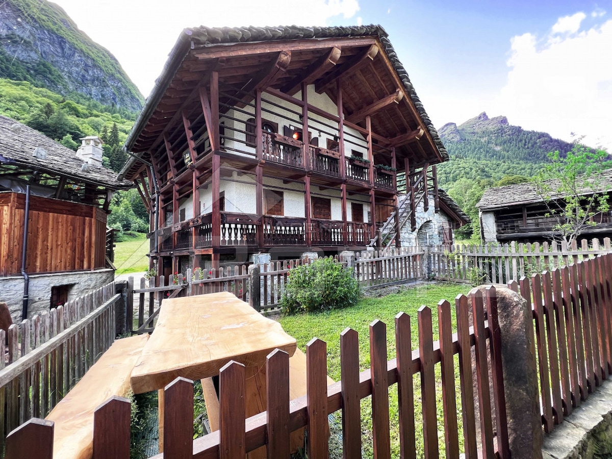 A charming wooden house with multiple levels is surrounded by lush greenery and mountain views. Exposed wooden beams and a balcony enhance the rustic appeal. A wooden table, set on a grassy area, is framed by a traditional wooden fence, inviting outdoor relaxation.