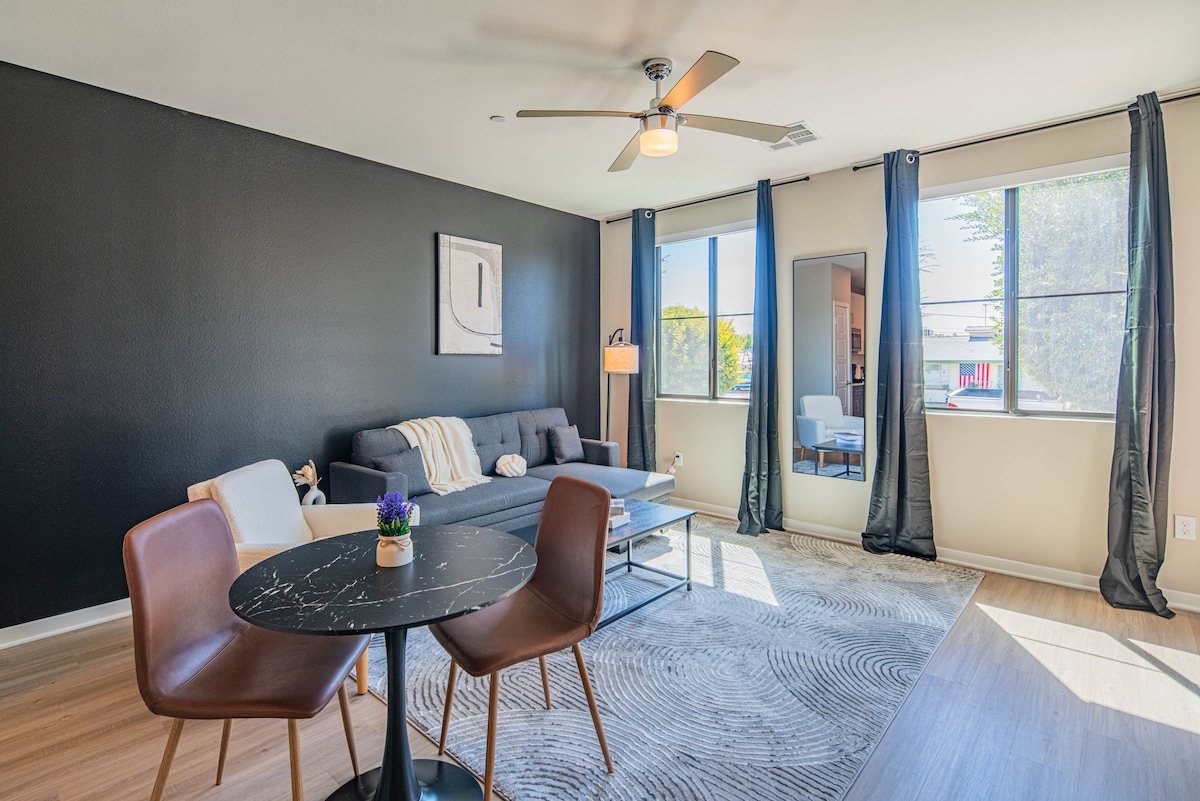 The living room features a modern design with a dark accent wall and large windows allowing natural light. A comfortable sofa is positioned with a coffee table in front, complemented by a pair of stylish chairs around a circular dining table.