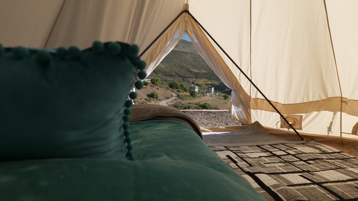 A tent interior features a plush teal pillow resting on a soft bed, with a textured rug placed underneath. A view of the surrounding landscape can be seen through the open tent flap, providing a glimpse of the outdoors.