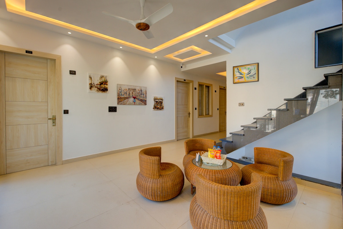 A modern living area features a series of round, textured chairs arranged around a small table. Soft, warm lighting accentuates the stylish decor, which includes framed artwork on the walls. A staircase leads to the upper level, while a television is mounted for entertainment.