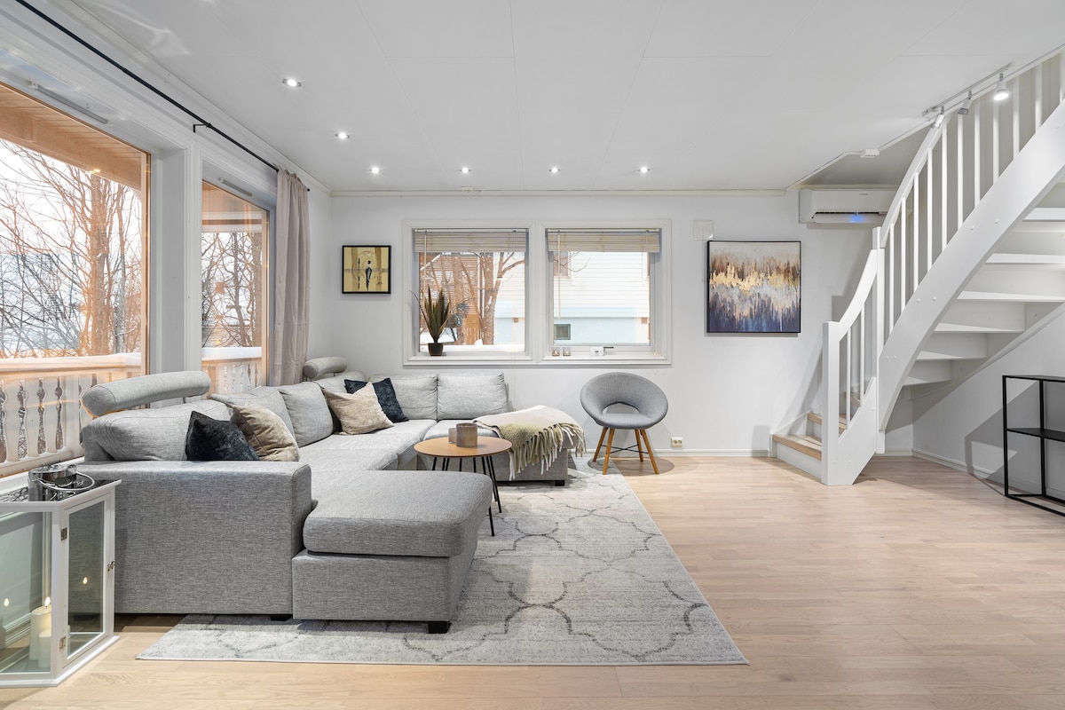 A spacious living area features a large, comfortable sofa and a round coffee table. Natural light filters through the windows, illuminating the soft color palette. Decorative wall art and a staircase add to the inviting ambiance of the space, which is equipped with a cozy chair.