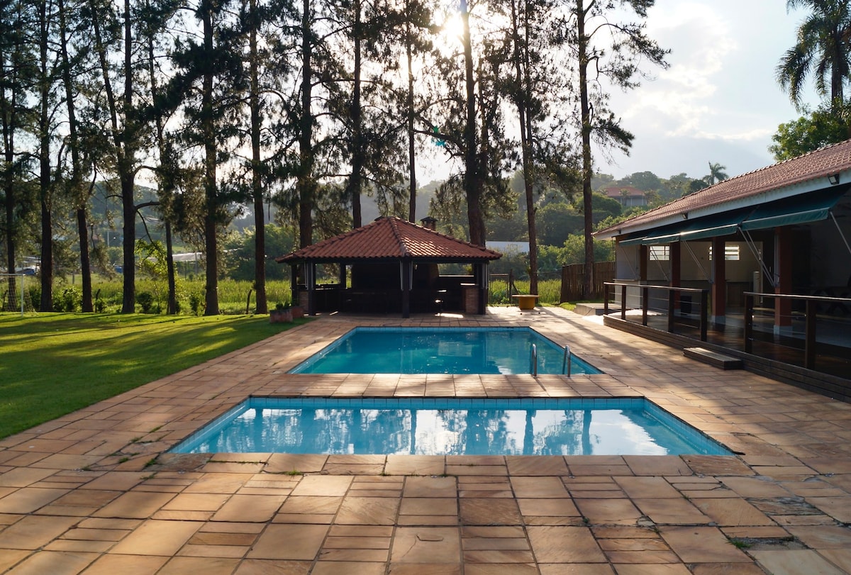 An outdoor area features two swimming pools, one large and one smaller, surrounded by a spacious tiled patio. A shaded gazebo with a thatched roof offers seating nearby. Lush greenery and tall trees are seen in the background, illuminated by soft sunlight.