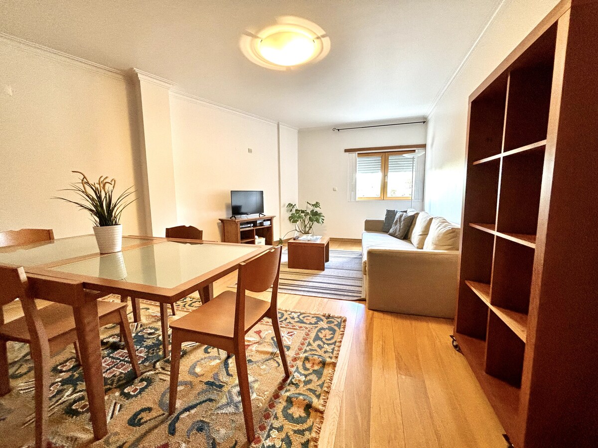 A light-filled living area features a spacious sofa and a wooden dining table surrounded by chairs. A television is set on a cabinet, while a decorative plant adds a touch of greenery. The wooden flooring complements the area, providing a warm ambiance.