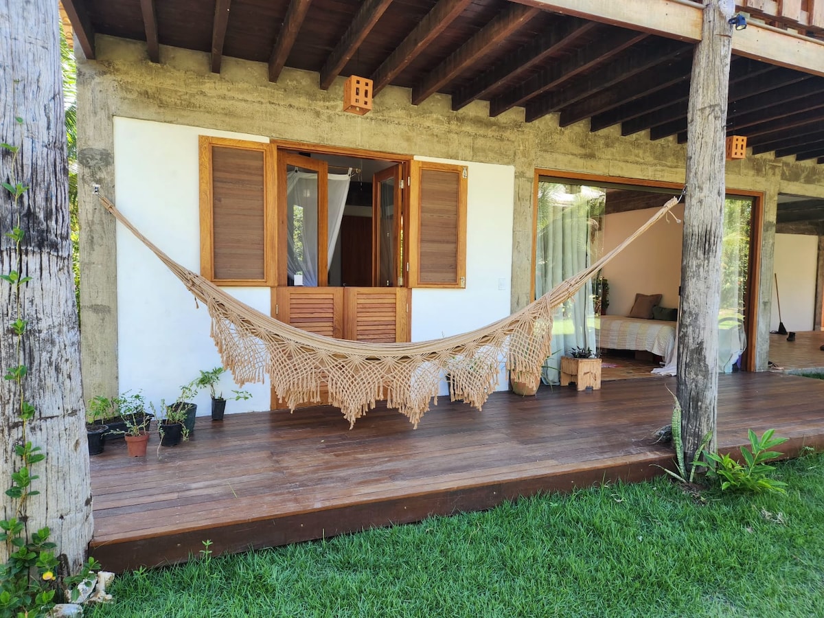 A wooden deck opens to a cozy outdoor area featuring a hammock suspended between two posts. Potted plants are arranged along the edges, while large glass doors lead into a well-lit indoor space. The textured walls provide a natural backdrop.