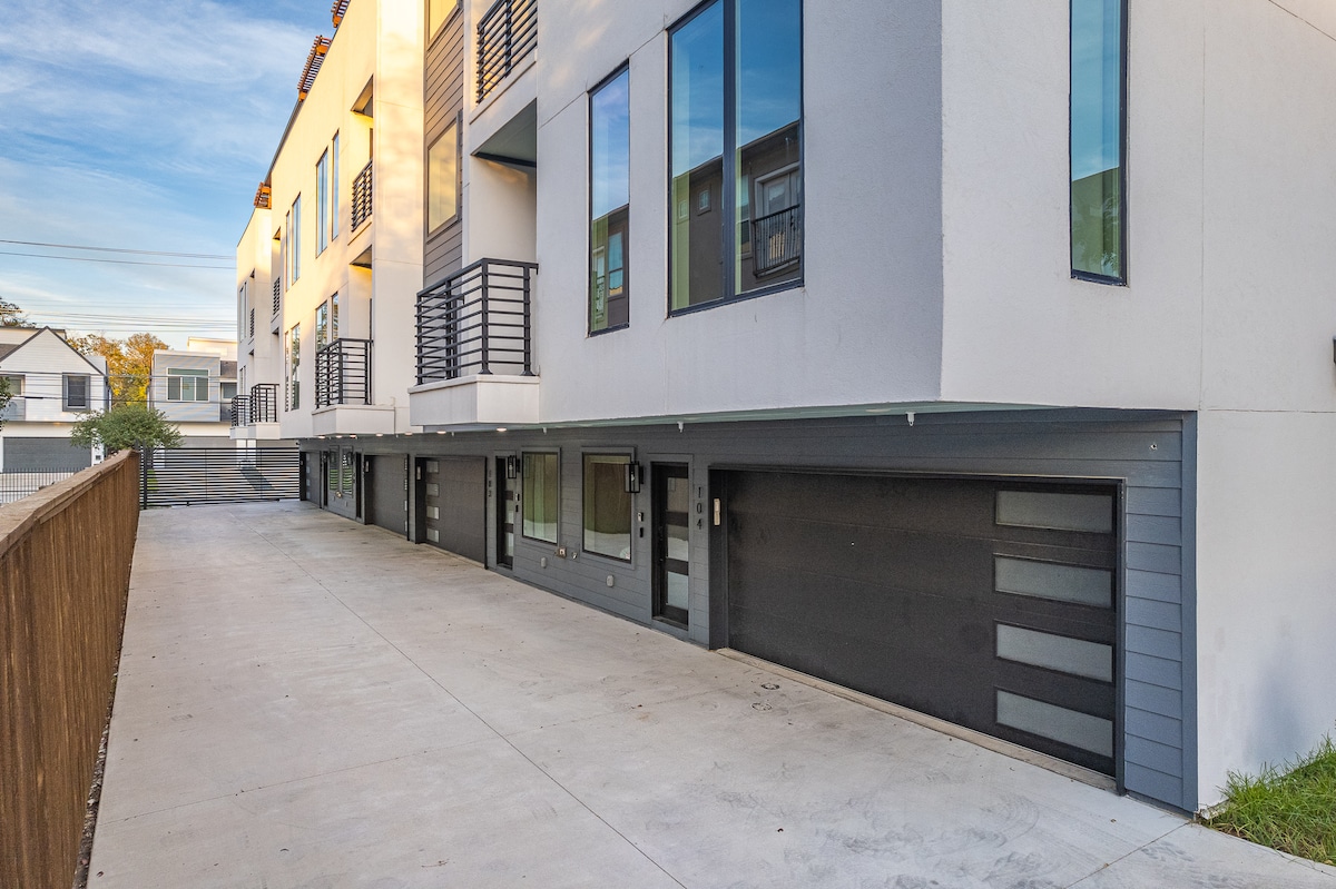 The exterior of the home features a modern architectural design with multiple large windows that allow natural light to fill the interior. A spacious driveway leads to two garage doors below, offering ample parking and access to the house. The surrounding area includes a landscaped yard.