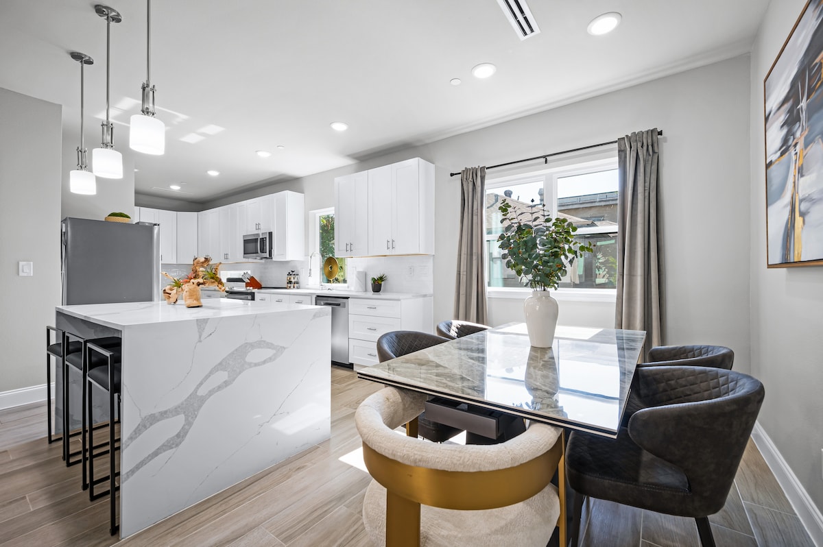 A modern kitchen and dining area features white cabinetry with stainless steel appliances. A marble island with bar seating complements a glass dining table surrounded by stylish chairs. Natural light filters through a window adorned with soft curtains, illuminating the space.
