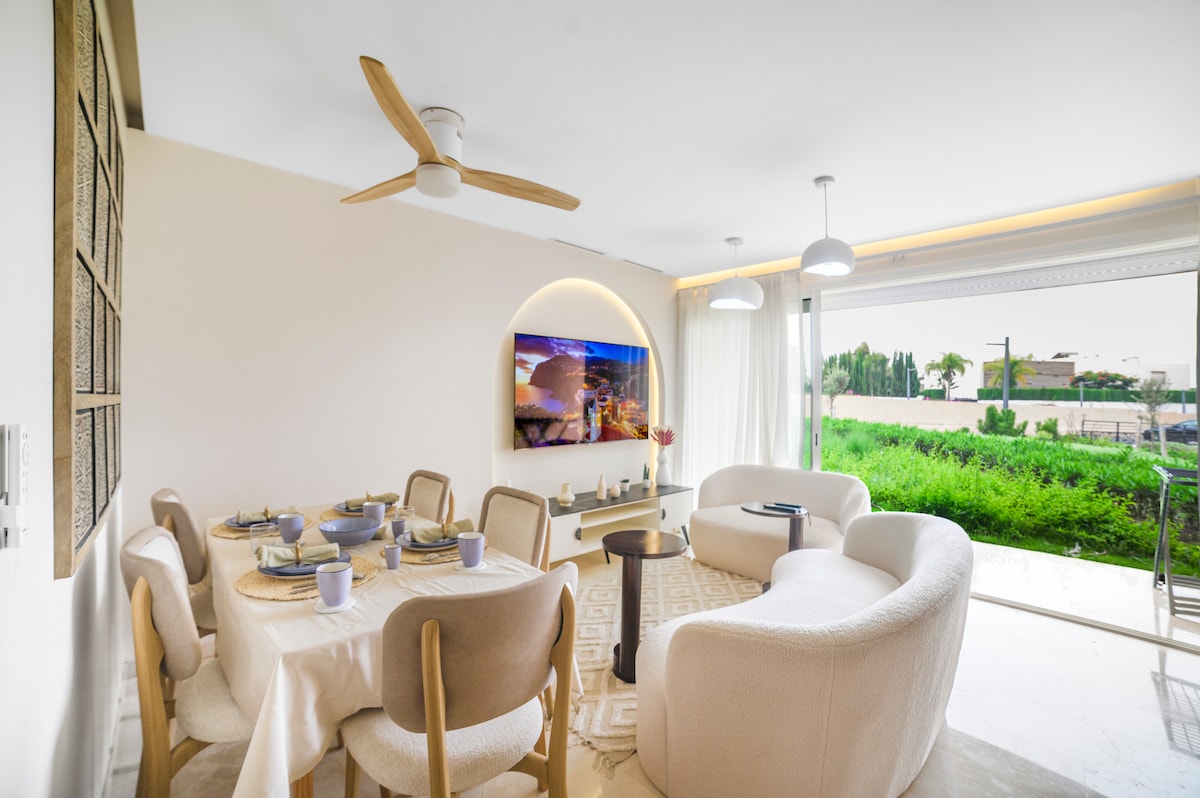 A bright and airy living area is furnished with a circular white sofa and a spacious dining table set for six. Natural light floods in through large windows, with greenery visible outside. A mounted TV is displayed on the wall, enhancing the modern ambiance.