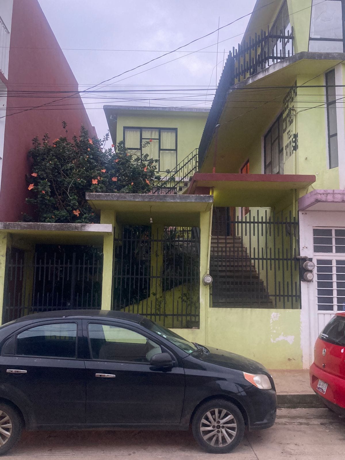 A two-story building features a vibrant green exterior, with large windows and a staircase leading to the upper floor. Flowering plants are visible near the entrance, and parked cars are seen in the foreground, providing a sense of accessibility to the space.
