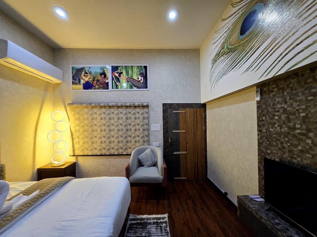 A well-furnished room features a spacious bed with clean linens and decorative pillows. A soft armchair is positioned near the door. The wall displays large art pieces and a vibrant peacock design, while the room is brightly lit with modern fixtures.
