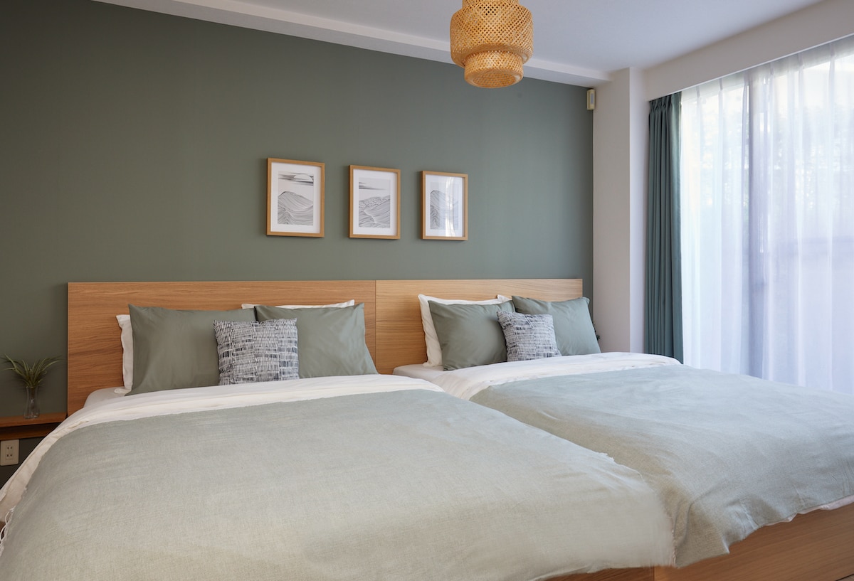 A bright bedroom features two spacious beds adorned with light green bedding. Each bed is accented by decorative cushions and a framed wall art trio above. Natural light filters through large windows, complemented by soft curtains, and a simple plant rests on the bedside table.