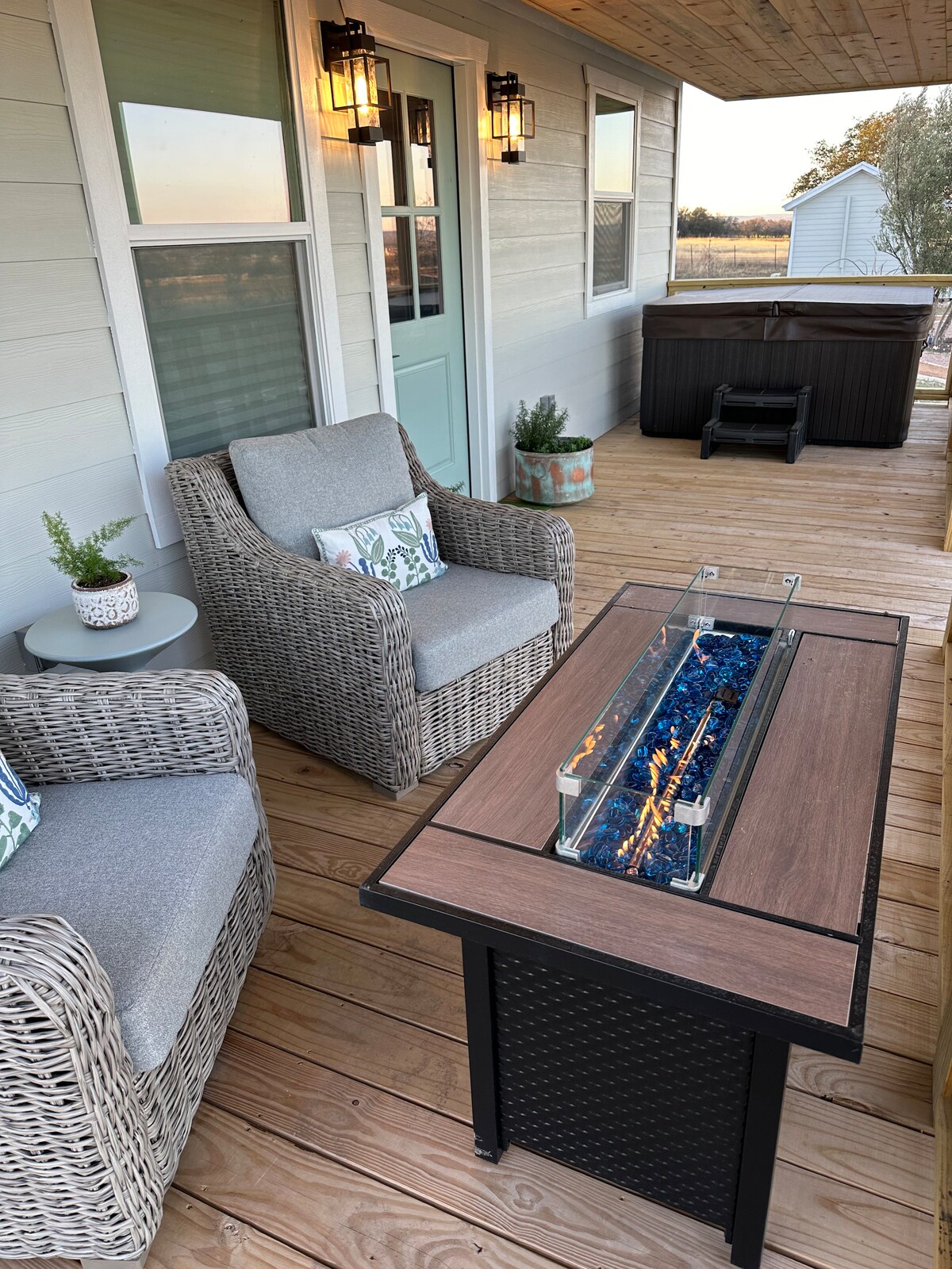 A cozy deck area features two cushioned wicker club chairs positioned around a modern fire table with a glass top. A hot tub is visible nearby, with a serene view of the surrounding landscape through an open door.